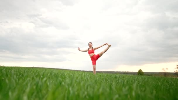 Woman in orange sports wear practicing yoga - utthita hasta padangushthasana in green field. Balance on one leg. fitness, sport, healthy lifestyle concept. — Αρχείο Βίντεο