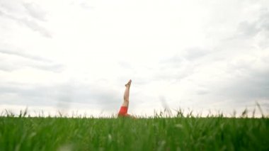 Strong woman in orange wear doing yoga Sarvangasana - shoulder stand, inverted asana in green field. Girl building strong core, focused and motivated.