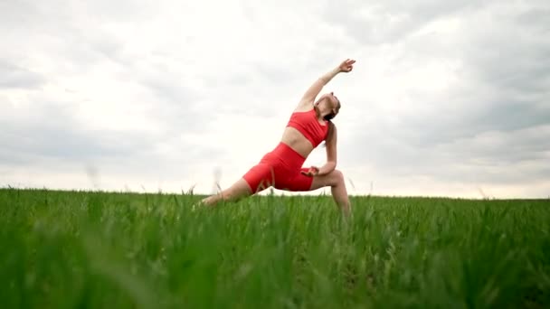 Sporty woman in orange costume practicing yoga in green fresh field, minimalist scene. Doing stretching asana with legs and arms. Healthy lifestyle, breathe deeply concept. — Stockvideo