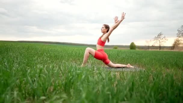 Turuncu kostümlü genç kadın yeşil alanda yoga yapıyor. Minimalist bir sahne. Asana 'yı bacak ve kollarla esnetiyorum. Sağlıklı yaşam tarzı, derin nefes al konsepti. — Stok video