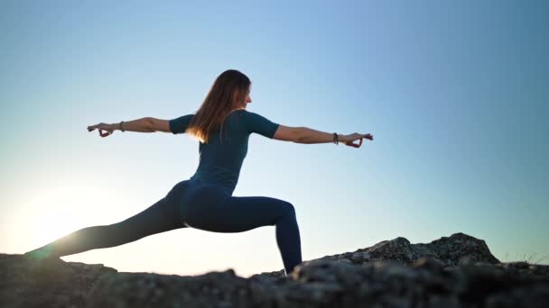 Vrouw doet yoga Virabhadrasana - krijger poseren op hoge heuvel, hemel achtergrond, zonsondergang tijd. Stretching, dagelijkse praktijk, gezonde levensstijl concept — Stockvideo