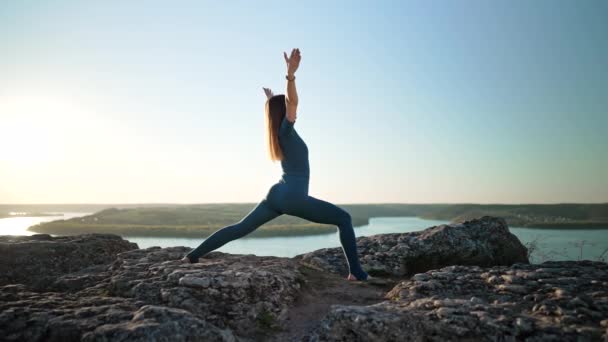 Sportieve vrouw in blauw kostuum die yoga beoefent op hoge rots boven water. Asana 's strekken, oefeningen met benen en armen. Gezonde levensstijl, diep inademen concept. — Stockvideo