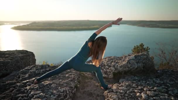 Αθλητική γυναίκα με μπλε στολή που κάνει γιόγκα σε ψηλό βράχο πάνω από το νερό. Κάνοντας διατάσεις asanas, σειρά ασκήσεων με τα πόδια και τα χέρια. Υγιεινός τρόπος ζωής, βαθιά αναπνοή έννοια. — Αρχείο Βίντεο