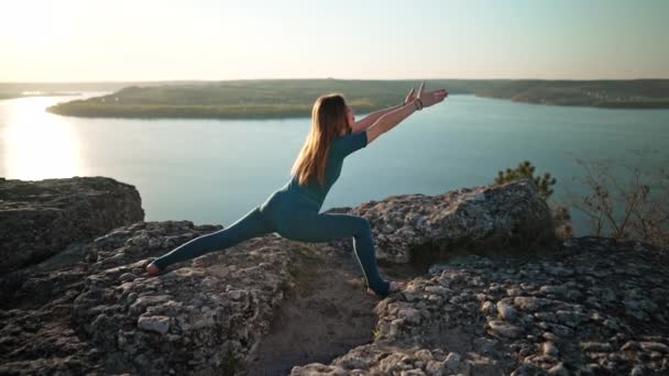 水の上の高い岩の上にヨガを練習青い衣装を着たスポーティ女性。ストレッチアサナ、足と腕の演習のシリーズを行う。健康的なライフスタイル深く呼吸する. — ストック動画