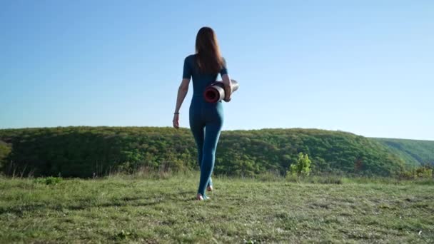 Egy fiatal nő sétál a reggeli jóga edzésre. Aktív lány fitnesz szőnyeggel, sportfelszereléssel. Zöld fű, természet háttér. Nyári nap. — Stock videók