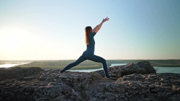 Sportowa kobieta w niebieskim stroju ćwicząca jogę na wysokiej skale nad wodą. Rozciąganie asan, seria ćwiczeń z nogami i ramionami. Zdrowy styl życia, oddychaj głęboko koncepcja. — Wideo stockowe