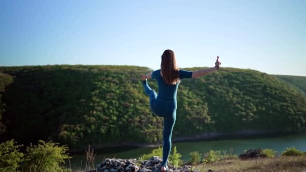 Femme en salopette bleue pratiquant le yoga utthita hasta padangushthasana sur une haute falaise au-dessus de l'eau. Équilibre sur une jambe. fitness, sport, mode de vie sain concept. — Video