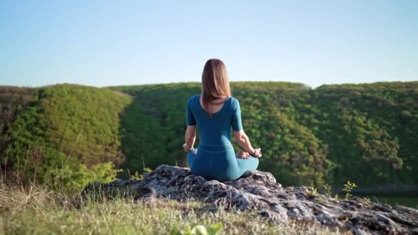 Skoncentrowana kobieta w lotosu pozuje medytując na wysokim klifie nad wodą. Koncepcja spokojnej jogi, religia, zen, spokojny umysł, praktyka na tle natury. — Wideo stockowe
