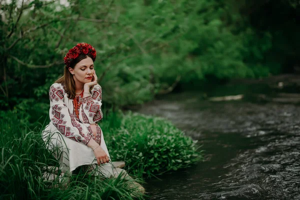Underbar ukrainska kvinna i traditionella broderi vyshyvanka klänning sitter nära floden på natur skog bakgrund. Ukraina, kultur, frihet, nationaldräkt. — Stockfoto