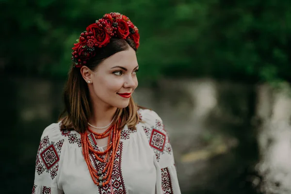 Csinos ukrán nő hagyományos hímzéses vyshyvanka ruhában és vörös virágkoszorúval a folyó mellett. Ukrajna szabadság, tavasz, nemzeti jelmez, győzelem a háborúban. — Stock Fotó