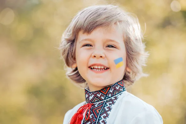 Um menino ucraniano sorridente com uma bandeira amarela azul na bochecha. Criança em tradicional bordado camisa vyshyvanka. Ucrânia, liberdade, traje nacional, vitória na guerra. — Fotografia de Stock