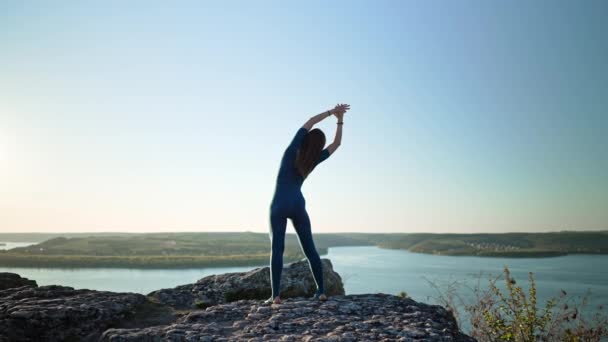 Egy jógi nő sziluettje, aki jógázni kezd a szikla tetején a magas hegyen. Vad természet, szabad ember. Szabadság, rugalmasság, mindennapi gyakorlat, testnyújtás — Stock videók