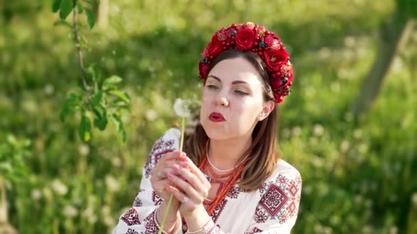 Femme ukrainienne en broderie traditionnelle vyshyvanka robe soufflant pissenlit dans un beau parc. Ukraine, liberté, printemps, costume national, mode authentique. — Video