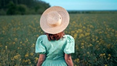 Çiçek açan kanola tarlasında poz veren çekici bir kadın. Hasır şapkalı, eski moda elbiseli zarif bir kız. Kolza tohumu çayırı, eski moda kıyafetler, bahar mevsimi. 4k görüntü