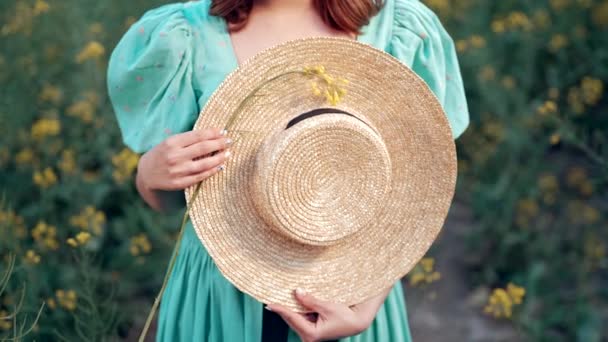 ブラックリボンで手をわらトレンディーな帽子を保持女性 夏には菜の花畑に立ち ヴィンテージブースターを転がす女の子 高品質4K映像 — ストック動画