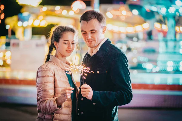 Millennial couple with sparkling bengal fire spending time together on date in amusement park at night. Lovers standing on Illuminated background. Celebration, anniversary. High quality photo