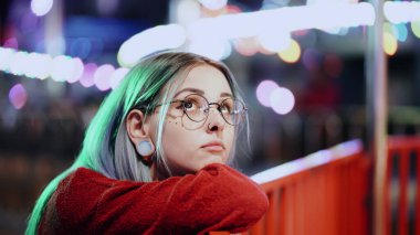 Hipster girl with blue dyed hair, golden sequins as freckles. Sad woman with nose piercing, transparent glasses, ears tunnels, unusual hairstyle stands in amusement night park. High quality photo