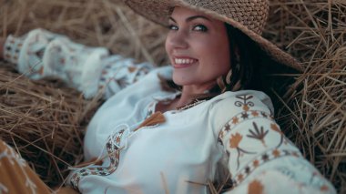 Pretty woman in straw hat and embroidered blouse smiling lying on hay in countryside at sunset. Rural nature, haystack, vacation, relax and harvest concept. High quality photo