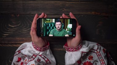 August 2022 - Kyiv, Ukraine. Watching online speech of President Volodymyr Zelenskiy on smartphone on wooden table background. News from front. russian aggression. High quality photo