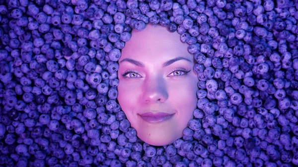 Smiling woman face in fresh ripe blueberries. Young attractive girl covered with acai berries. Lady with bright makeup. enjoying organic bilberry plant. Diet, antioxidant, vegan food. High quality