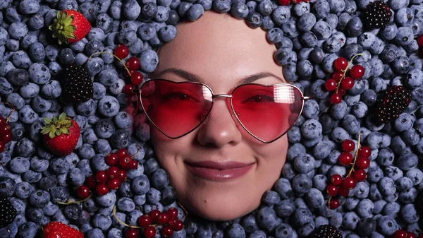 Happy woman face in eyewear fresh ripe berries - blueberries, strawberries, currant. Young girl covered with blackberry. Lady enjoying organic bilberry plant. Diet, antioxidant, healthy vegan food