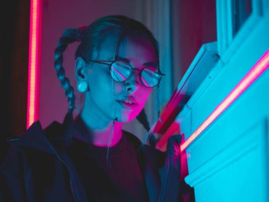 Şehrin gece parlayan pembe ve mavi neon ışıkları yakınında alışılmadık saç modeli genç güzel kız. Mavi boyalı saç örgü içinde. Ciddi hipster genç gözlük ve güzel lensler.