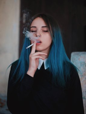 Emo girl smoking cigarette. Young student or pupil with blue colorful dyed hair, hat, piercing,lenses,ears tunnels and unusual hairstyle stands on black background.