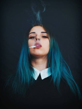 Emo girl smoking cigarette. Young student or pupil with blue colorful dyed hair, hat, piercing,lenses,ears tunnels and unusual hairstyle stands on black background.