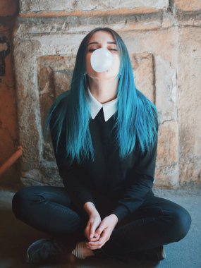 Cute gothic model blows bubblegum. Street punk or hipster woman student with blue colorful dyed hair, piercing,lenses,ears tunnels and unusual hairstyle in backyard.