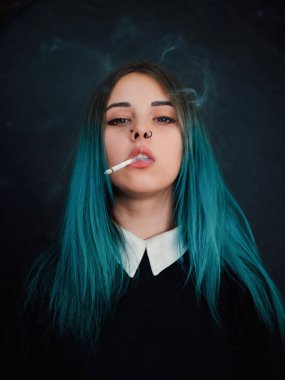 Emo girl smoking cigarette. Young student or pupil with blue colorful dyed hair, hat, piercing,lenses,ears tunnels and unusual hairstyle stands on black background.