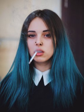 Emo girl smoking cigarette. Young student or pupil with blue colorful dyed hair, hat, piercing,lenses,ears tunnels and unusual hairstyle stands on black background.