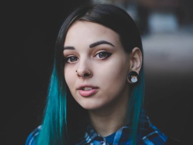 Street punk or hipster girl with blue dyed hair. Woman with piercing in nose, violet lenses, ears tunnels and unusual hairstyle stands in city.