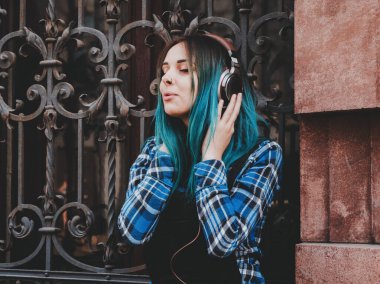 Street hipster girl listening music with brown headphones. Teen girl with blue dyed hair,piercing in nose,violet lenses,unusual hairstyle sitting near historic building.