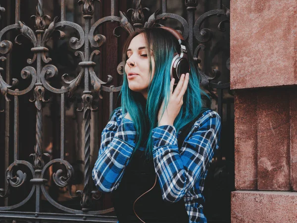 Street hipster girl listening music with brown headphones. Teen girl with blue dyed hair,piercing in nose,violet lenses,unusual hairstyle sitting near historic building.