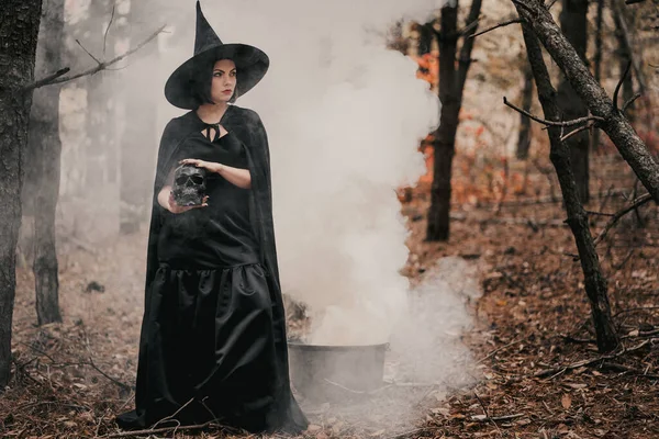 Black witch holding skull in hands, stroking it on autumn forest background. Horror, halloween, cosplay holiday, magic concept. High quality photo