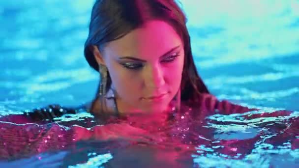 ネオン輝く色の光の下でプールの水の中でグラマラスセクシーな女性 パーティー 夜の時間を楽しんでドレスを輝いて魅力的な女の子 ポージングモデル インフルエンサーとして働く 高品質4K映像 — ストック動画