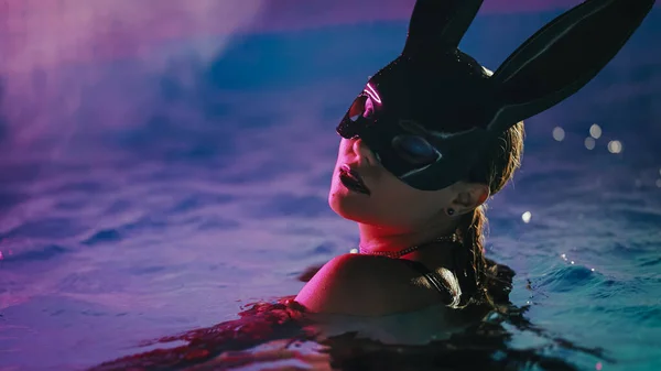 革のBdsmマスクの官能的な女性はネオンカラーの光の下でプールの水で魅惑的に移動します ハロウィンパーティー 大人のためのナイトゲームを楽しむ魅力的なヒヨコマスケードウサギ — ストック写真