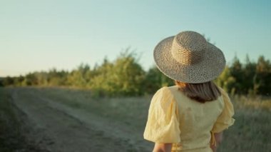 Tanımlanamayan bir kadın, retro elbiseli, ayçiçeği tarlasının yakınında tek başına yürüyor. Klasik moda, inanılmaz macera, kırsal alan, kırsal alan, doğal yaşam tarzı. Yüksek kalite 4k görüntü