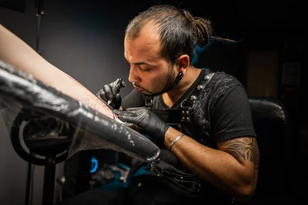 Portrait of man tattoo master with dreadlocks showing process of creation tattoo on female body under lamp light. Professional artist working in salon. High quality photo