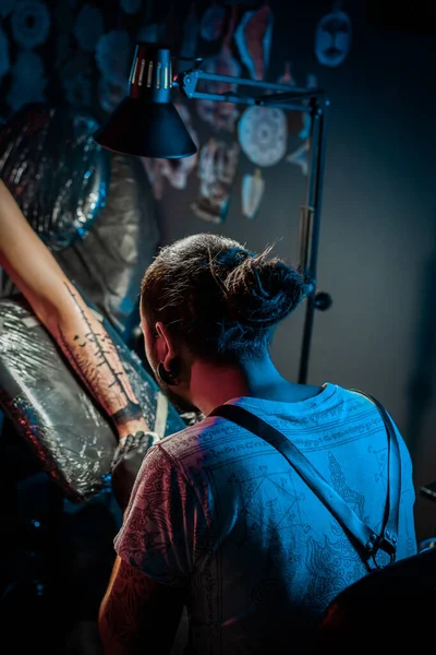 ランプの光の下で女性の体に創造の入れ墨のプロセスを示すドレッドロックと男の入れ墨のマスターの肖像 サロンで働くプロのアーティスト 高品質の写真 — ストック写真