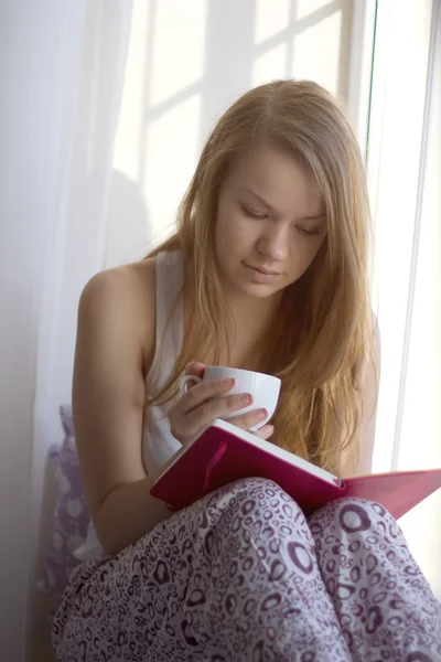 Flicka på fönsterbrädan med en kopp läser bok — Stockfoto