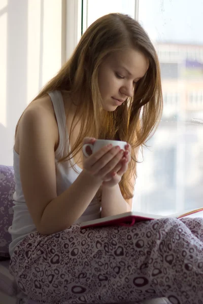Flicka på fönsterbrädan med en kopp läser bok — Stockfoto