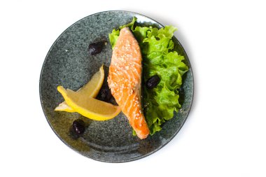 Beyaz arka planda bir dilim limon, marul ve zeytinle servis edilen somon bifteği.