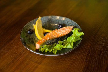 Fırında somon bifteği, bir dilim limon, marul ve zeytin ile servis edilmiş kahverengi ahşap masa arka planında.