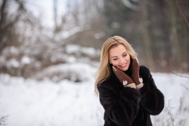 Kış karlı bir ormanın arka planında gülümseyen güzel bir sarışın kız sıcak kahverengi eldivenlerle yüzünü ısıtıyor.