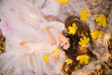 Açık bir günde, uzun siyah saçlı, düğün kabarık şifon elbiseli bir kız düşen sonbahar akçaağaç yapraklarının arasında uzanıyor.