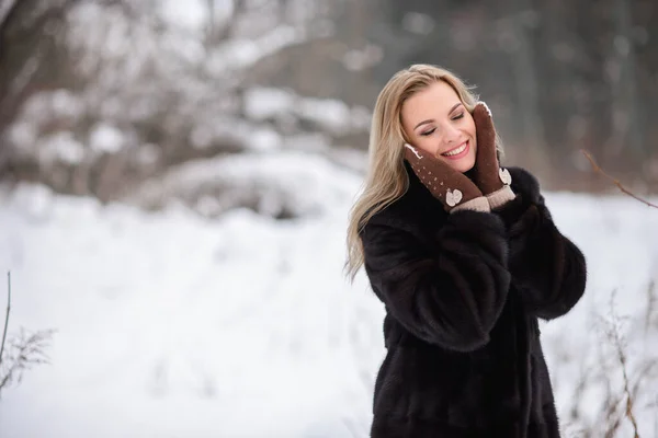 冬の雪の森の背景にある美しい笑顔ブロンドの女の子暖かい茶色の手袋で彼女の顔を暖めます — ストック写真