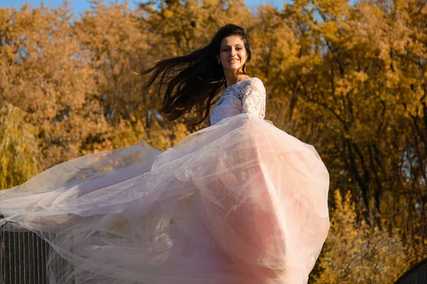 a girl with long dark hair in a lush pale pink ball gown in autumn on a sunny day, chiffon fabric flies from a gust of wind
