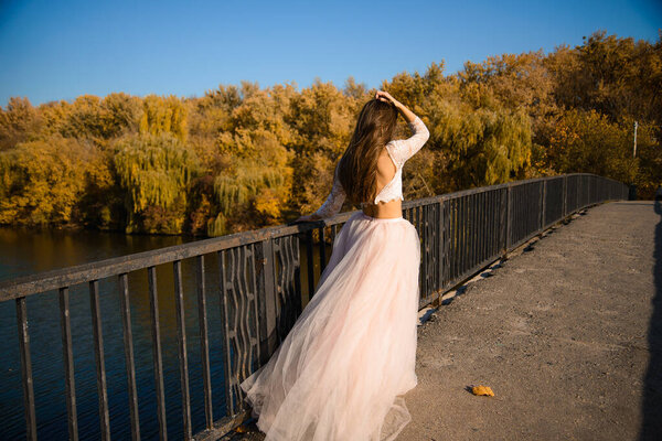 A girl with long dark hair in a lush pale pink ball gown in autumn on a sunny windy day, walks along the bridge