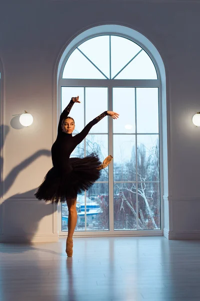 漂亮的芭蕾舞演员 身穿黑色紧身衣和短裙 背靠大窗户 黑暗房间里有拱门 踮起脚尖 — 图库照片